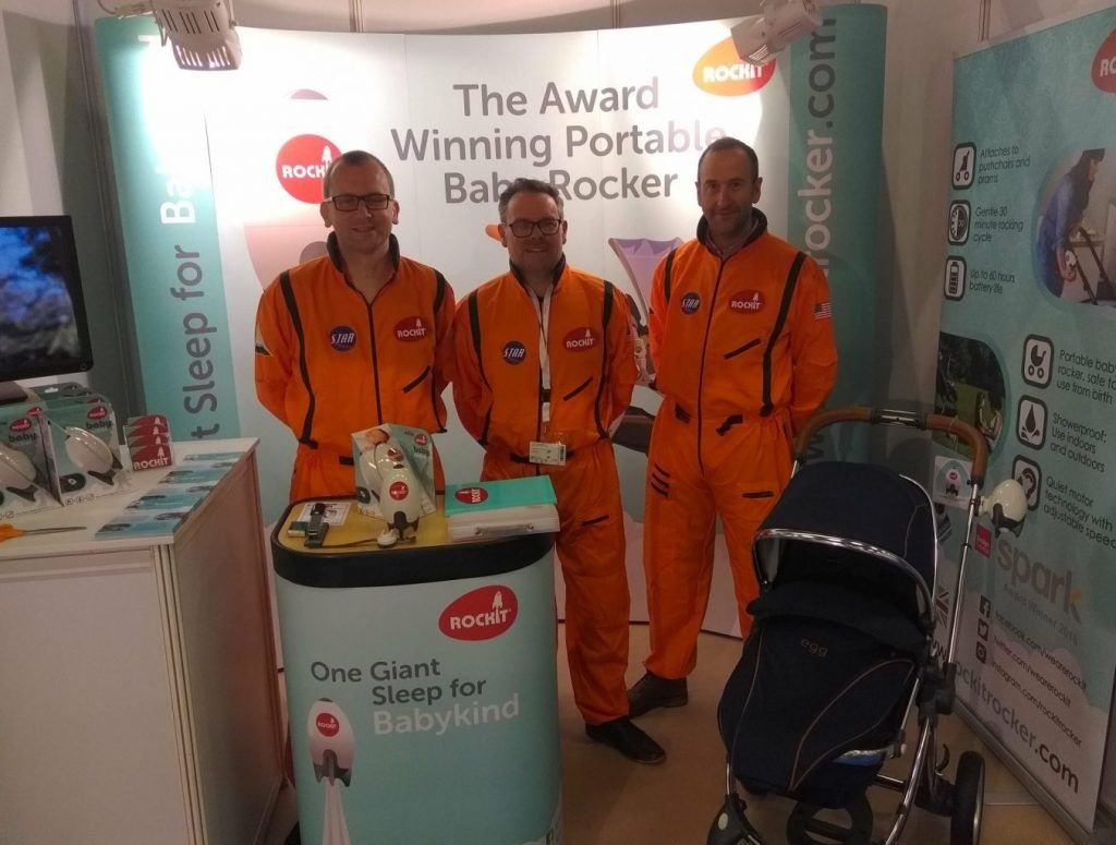 Three men dressed in orange space suits at an exhibition stand. The exhibition shows a display of the portable baby rocker called Rockit and includes the logo, product samples and information about the product
