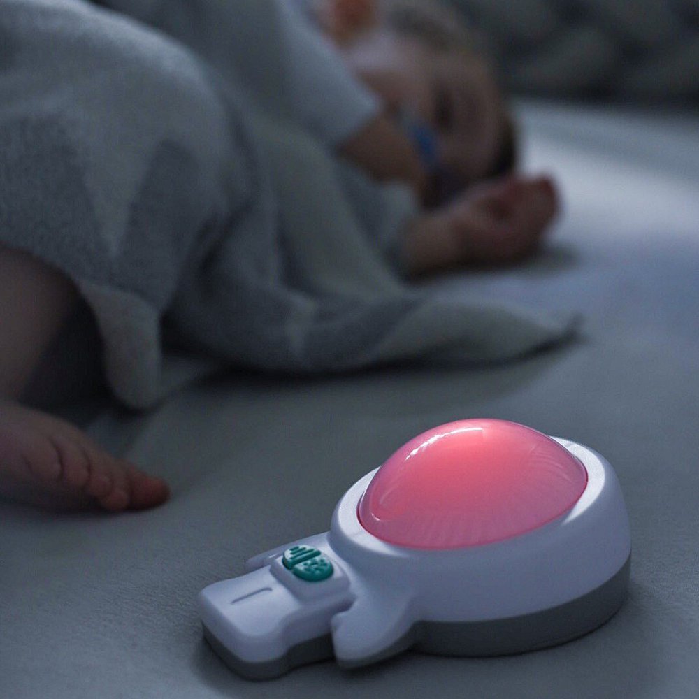 Close up of a baby soothing device called Zed with a sleeping baby in the background on the same mattress.
