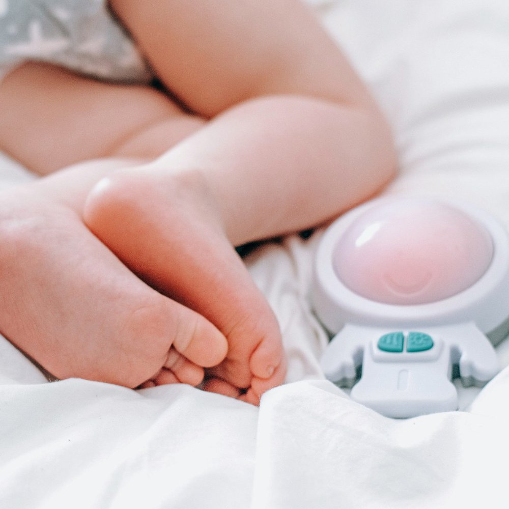 Close up of a babies legs and feet on a while sheet and a soothing device called Zed next to them